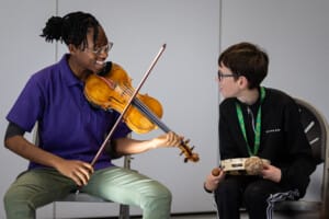 Young Associate Àánú working with our Family Orchestra in Exeter. She is seated playing the violin with a participant and is wearing a purple t-shirt