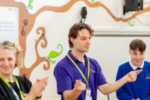 Young Associate, Ed, at Linwood School. He is leading an activity alongside Associate Musician Sam Mason. He is wearing a purple t shirt and standing alongside a student and Sam Mason