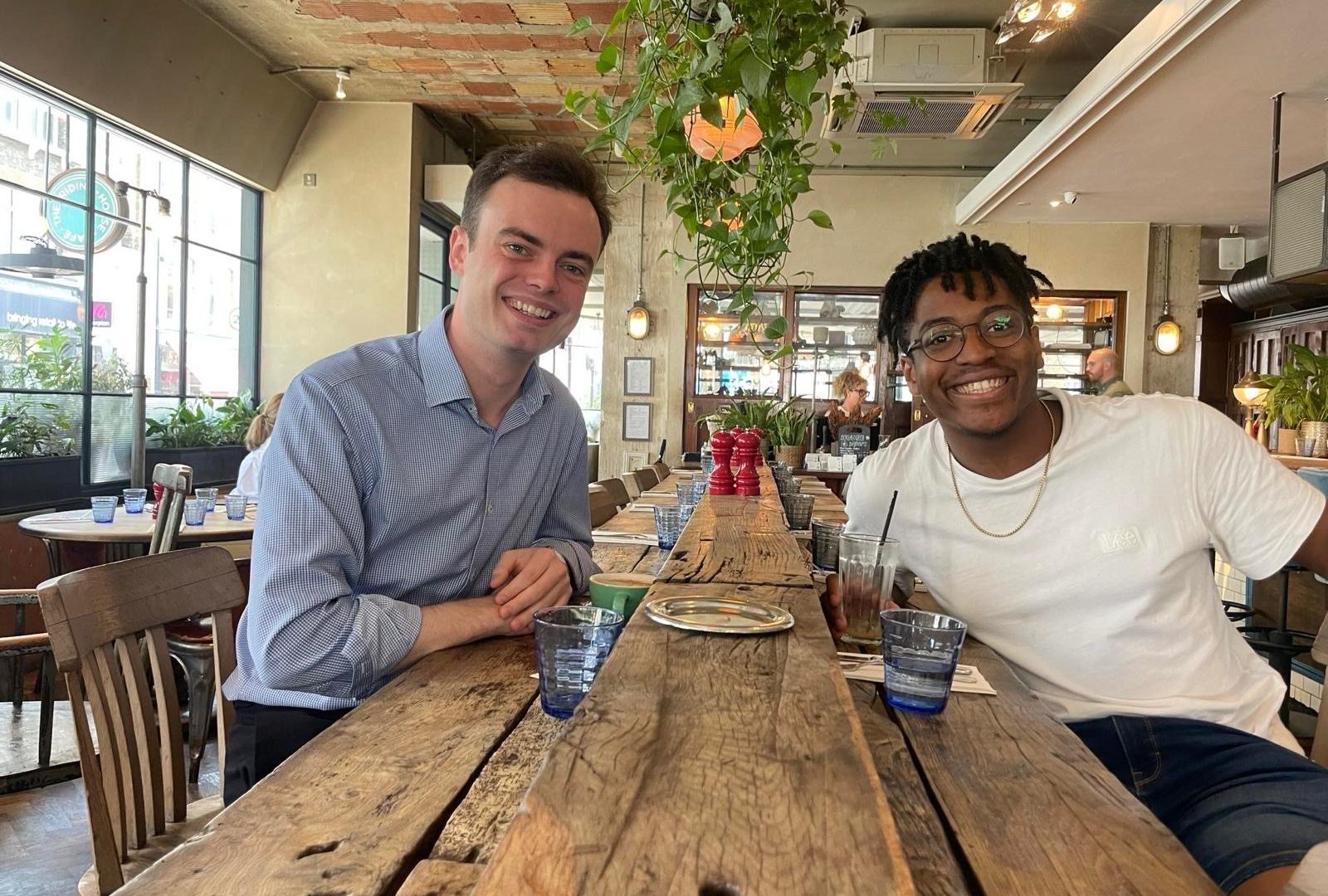 Previous BSO Assistant Conductor, Tom Fetherstonhaugh and new BSO Calleva Assistant Conductor Enyi Okpara sitting in a cafe and smiling a the camera.