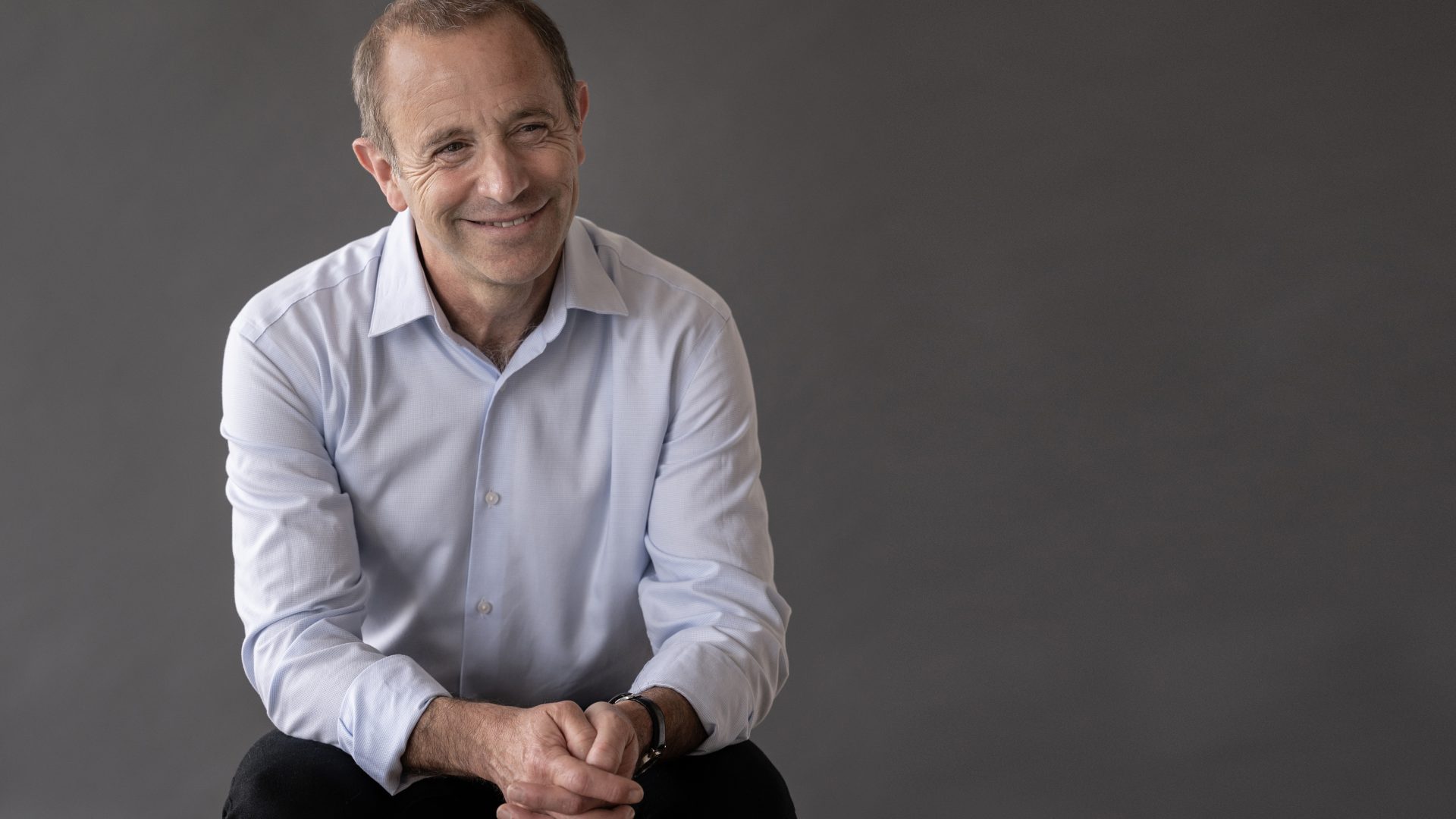 Conductor Mark Wigglesworth sits with hands together and smiling towards the right-hand side.