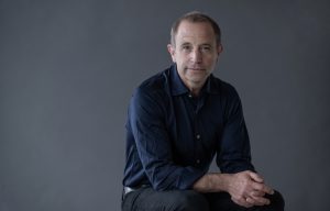 Conductor Mark Wigglesworth sits against grey background, wearing a dark blue shirt