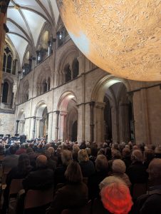 Full church nave shown with orchestra in background. Luke Jerram's 'Mars' art installation hangs from the ceiling