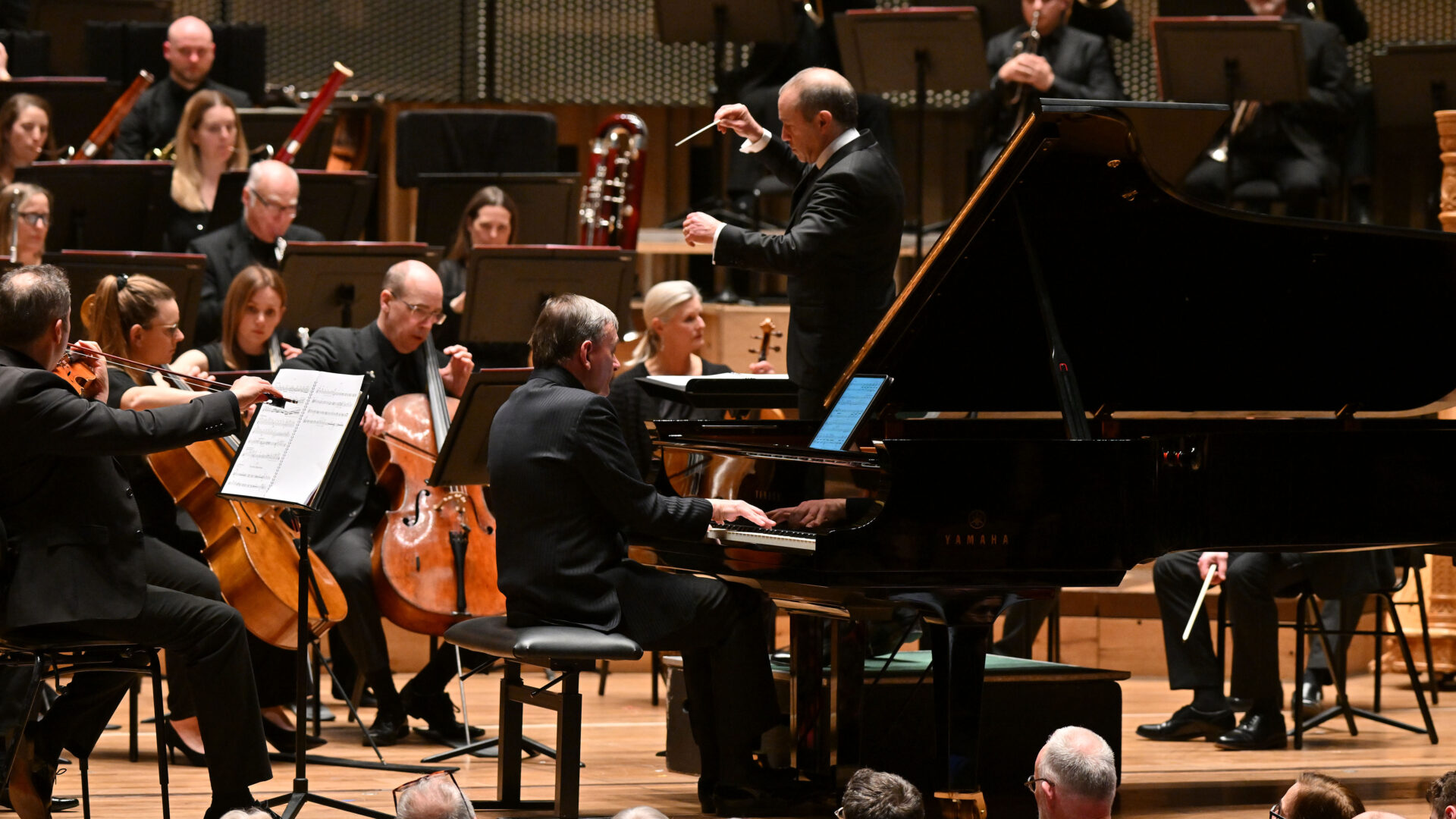 At the Bristol Beacon, renowned conductor Mark Wigglesworth leads the Bournemouth Symphony Orchestra, with a pianist gracefully playing a grand piano in the foreground. The audience is captivated by the enchanting performance.