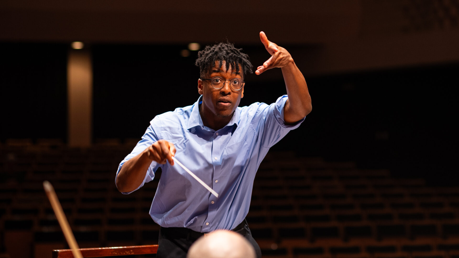 Conductor Enyi Okpara conducting and leaning over the podium facing the camera with baton in hand and left hand extended upwards.