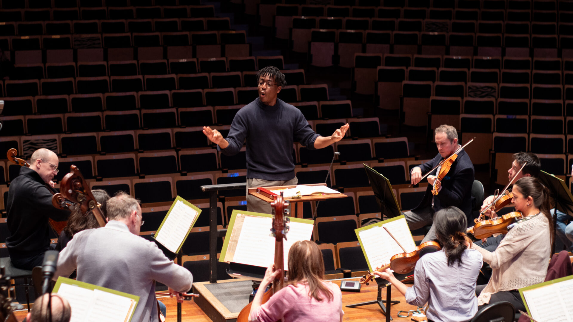Condcutor Enyi Okpara in a dark sweater faces the camera and looks at the cellos of an orchestra as he conducts with his hand and a baton.