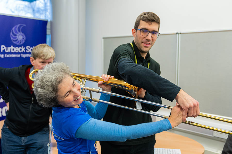 Musician in blue t-shirt has a touch tour of a trombone to sense how it feels to hold and play. They are guided by a trombonist, who is guiding them to feel the movement of the instrument. 