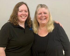 Two ladies who are part of BSO Voices stand with arms around each other wearing their BSO Voices uniform of black tops with logo
