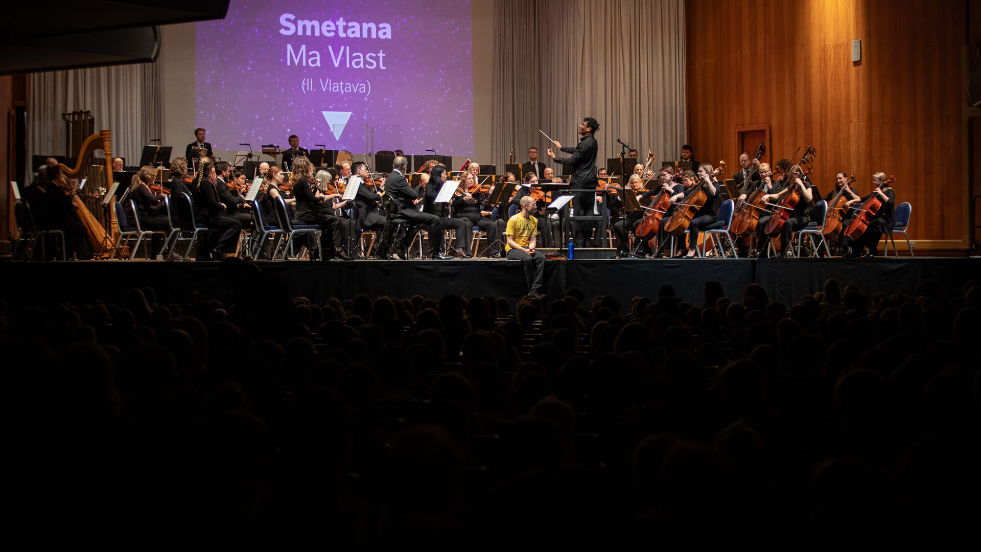 Orchestra and presenter perform a concert on stage to audience of hundreds of children
