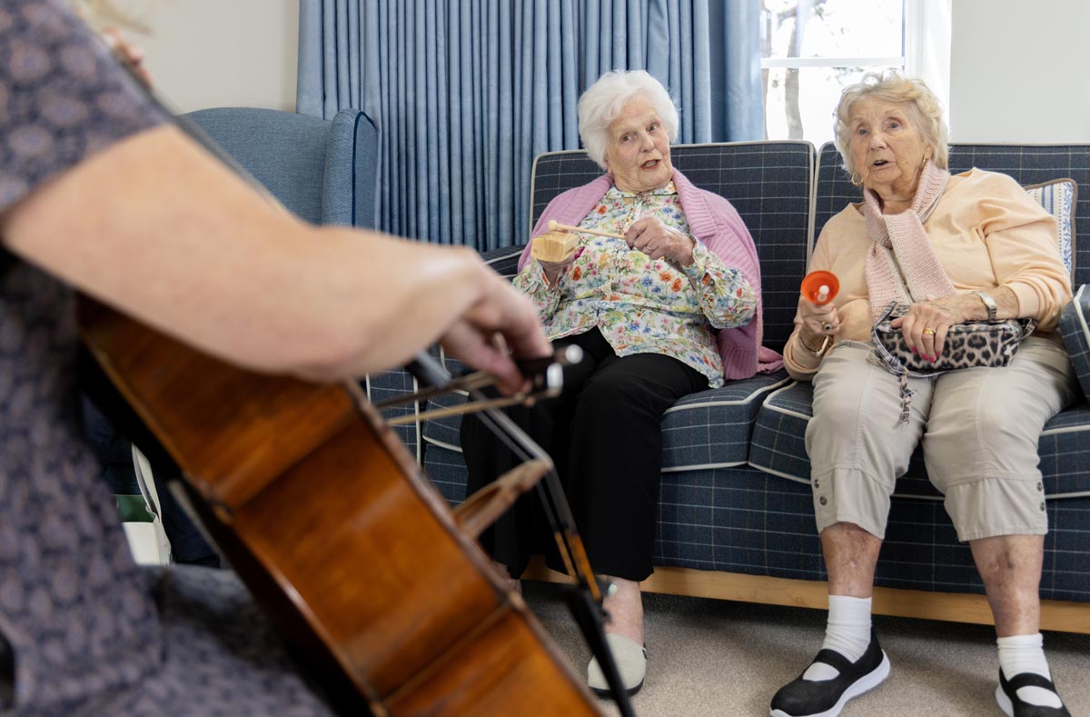 Two elderly ladies sit on a sofa with musical instruments facing a cellist