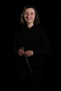 Michal Oren smiles to camera with conductors baton in hand. She is wearing a black outfit against a black background.