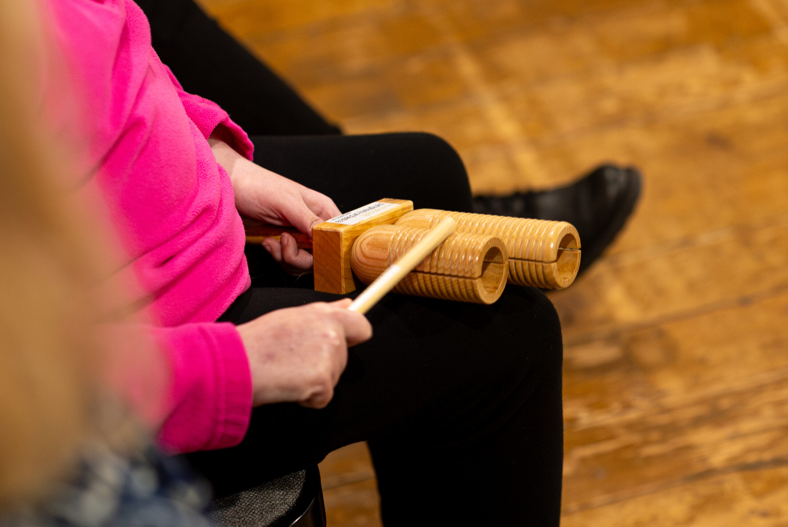 Person uses their hands to play a small wooden percussion instrument, holding a beater in their right hand.