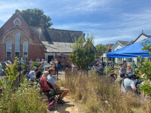 Musicians from Bournemouth Community Orchestra play music at The Old School House's summer celebration outside old building, and under a blue marquee. The sky is blue, sun is shining and there are lots of trees and plants. People sit on chairs and watch them play.