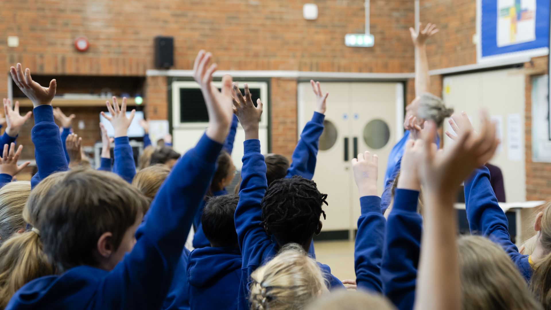 Schoolchildren in blue uniforms raise their hands in a classroom or school hall during a lesson.