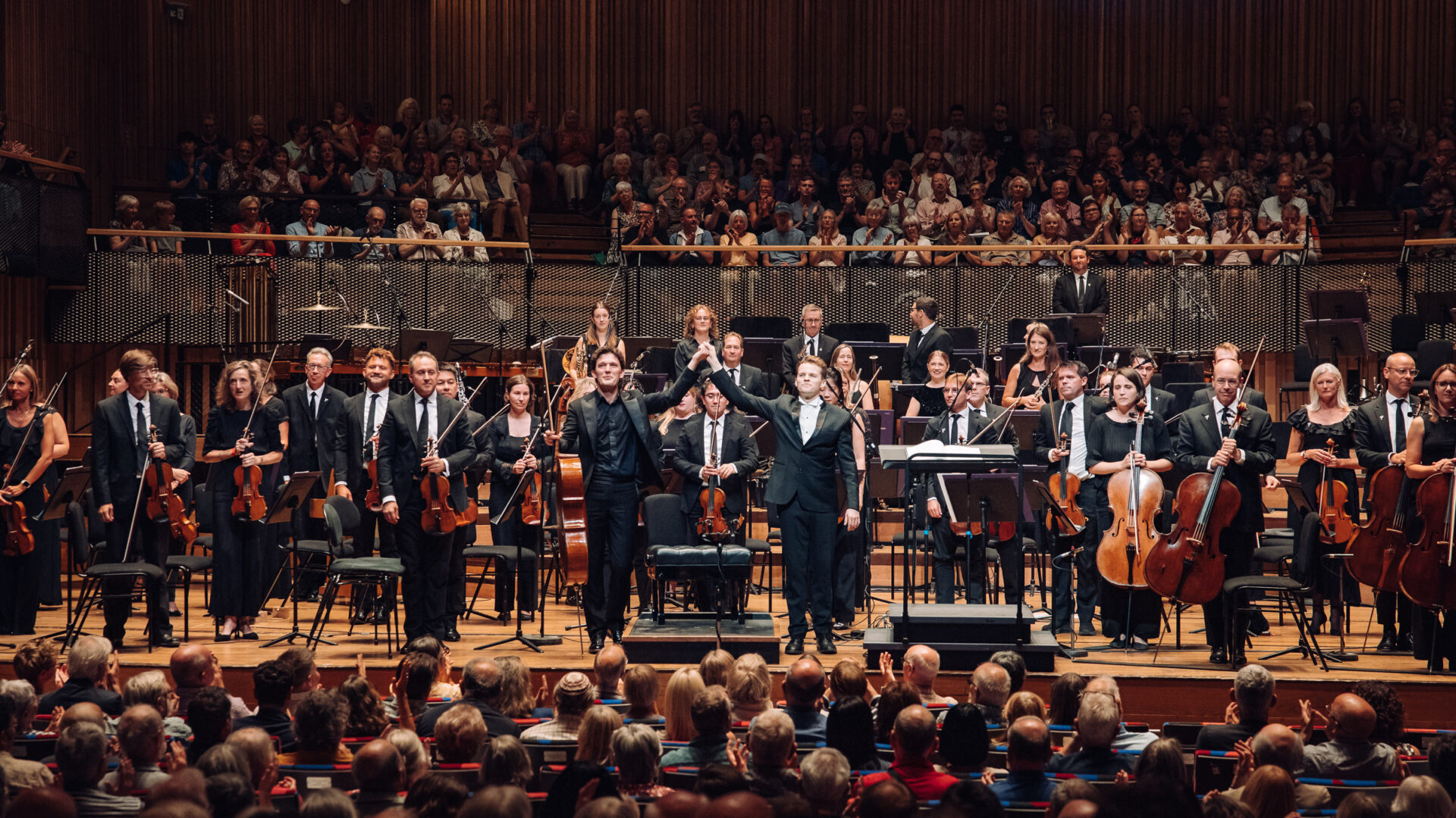 BSO on stage at Bristol Beacon following their BBC Proms performance, standing up as the crowd applauds. Conductor Adam Hickox and cellist Maximilian Hurnung raise their hands together.